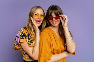 dichtbij omhoog portret van twee elegant Dames in zonnebril en modieus zomer kleren poseren Aan Purper bakgrond in studio. foto