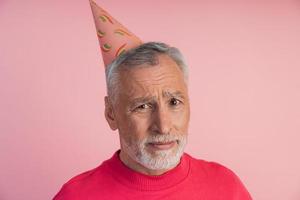 verdrietig, boos senior man in een feestelijke hoed op een roze achtergrond. foto