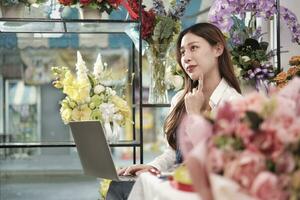 een jong Aziatisch vrouw bloemist en mkb ondernemer werken met laptop en denkt van inspirerend ideeën voor bloemen regelingen in kleurrijk bloem winkel met mooi bloesems, en een e-commerce bedrijf. foto
