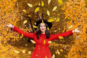 glimlachen vrouw in een zwart hoed en met rood lippenstift het werpen geel bladeren omhoog in de herfst Woud park foto