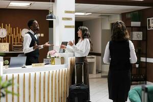 Kaukasisch senior vrouw ontvangt warm bijstand van Afrikaanse Amerikaans personeel Bij hotel voorkant bureau. mannetje receptioniste cheques in ouderen vrouw klant met bagage. ideaal voor vakanties. foto