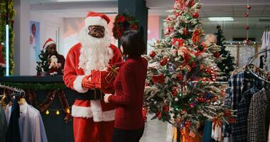 Afrikaanse Amerikaans werknemer acteren net zo de kerstman claus zittend naar beneden De volgende naar Kerstmis boom met Aziatisch vrouw in Kerstmis overladen kleding op te slaan. mode winkel arbeider aanbieden geschenk naar klant foto