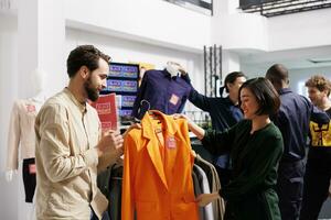 gelukkig verschillend paar boodschappen doen Aan zwart vrijdag in kleding op te slaan, op zoek Bij oranje formeel jasje met rood uitverkoop label en bespreken prijs. man helpen vrouw naar Kiezen kleren gedurende seizoensgebonden verkoop foto