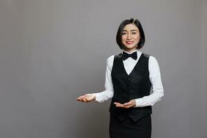 glimlachen Aziatisch vrouw serveerster vervelend restaurant uniform tonen met handen en groet gasten portret. professioneel receptionisten gastvrij klanten en op zoek Bij camera foto