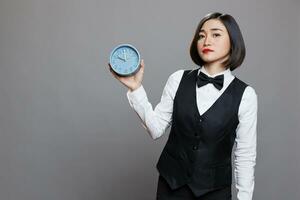 hotel vrouw receptioniste Holding retro alarm klok en op zoek Bij camera met echt gelaats uitdrukking. jong Aziatisch serveerster vervelend uniform tonen tijd beheer concept portret foto