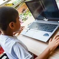 weinig jongen zittend Bij tafel gebruik makend van laptop voor online klasse in rang 1, kind aan het studeren Aan laptop van huis voor afstand aan het leren online opleiding, school- jongen kinderen levensstijl concept foto
