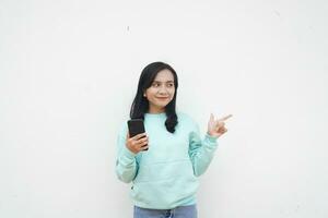 portret Indonesisch mooi vrouw vervelend licht blauw trui richten naar wit achtergrond houden mobiel telefoon met gelukkig gezicht foto