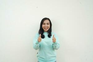 portret Indonesisch mooi vrouw vervelend licht blauw trui met glimlachen gelukkig gezicht en duim omhoog, geven loven, mee eens zijn, geïsoleerd Aan wit achtergrond foto