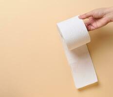een vrouw hand- houdt een rollen van wit toilet papier tegen een beige achtergrond foto