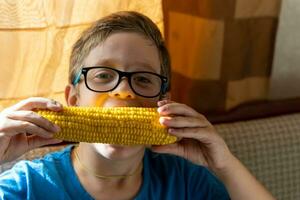 gelukkig schattig Kaukasisch jongen met bril aan het eten gekookt maïs Aan de maïskolf foto