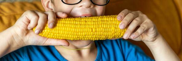 een gelukkig schattig jongen is aan het eten gekookt maïs Aan de maïskolf foto