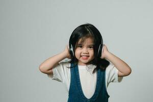 portret van ontspannen schattig weinig Aziatisch meisje kind grappig glimlachen en lachend vervelend wit koptelefoon, houdt handen Aan haar oren, poseren en glimlachen genieten van geïsoleerd over- grijs achtergrond. foto