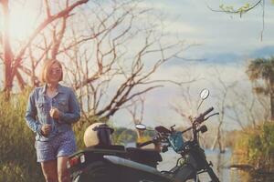 mooi vrouw toothy glimlachen met geluk staand naast klein enduro motorfiets Bij oerwoud veld- foto