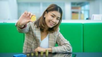 mooi Aziatisch bedrijf vrouw is zittend met een hand- Holding een gouden bitcoin verheven in voorkant van terwijl Daar een stapel van munten een rekenmachine en een tablet Aan zwart tafel, focas Aan goud bitcoins. foto