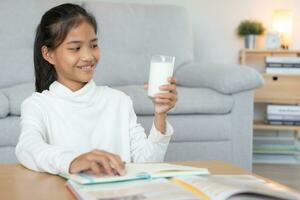 kinderen Dames drinken melk naar voeden de lichaam en voeden de brein. Aziatisch jong weinig meisje leren Bij huis. meisje gelukkig drinken melk en lezen een boek voor examen, thuisschool. opleiding, vitamines, ontwikkeling foto