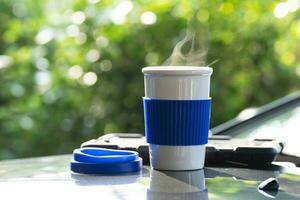 heet keramisch wit koffie kop met rook geplaatst buiten de auto dashboard Bij zonsopkomst in de ochtend, in een natuurlijk achtergrond. heet koffie drinken concept, selectief focus, zacht focus. foto