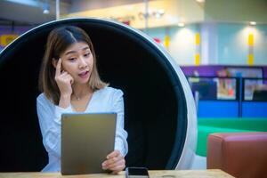 mooi Aziatisch bedrijf vrouw zittend Aan een modern ronde stoel Holding een tablet met een verrast uitdrukking, digitaal marketing. foto