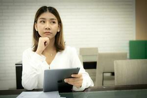 mooi Aziatisch bedrijf vrouw is zittend doen alsof naar denken in kopiëren ruimte met een twijfelachtig uitdrukking en Holding tablet Aan haar bureau, benadrukt kantoor meisje concept. foto