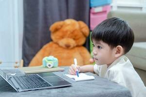 Aziatisch jongen nemen aantekeningen Aan papier terwijl aan het studeren online Aan laptop. foto