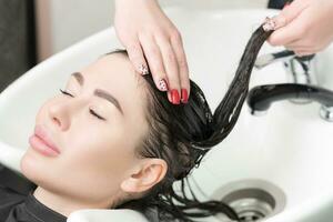 Haarstylisten handen wassen lang haar- van brunette vrouw met shampoo in speciaal wastafel voor shamponeren foto