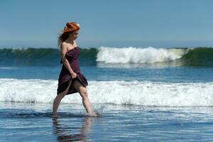 gelukkig roodharige vrouw wandelen enkel diep in water van oceaan golven Aan strand gedurende zomer vakantie foto