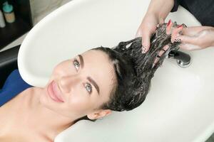 Haarstylisten handen wassen lang haar- van brunette vrouw met shampoo in speciaal wastafel voor shamponeren foto