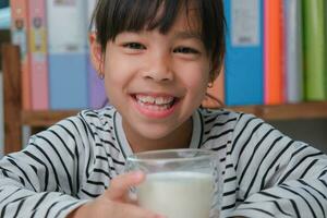 schattig Aziatisch meisje drinken een glas van melk Bij huis in leven kamer. weinig meisje drinken melk in de ochtend- voordat gaan naar school. gezond voedsel in jeugd. foto