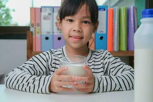 schattig Aziatisch meisje drinken een glas van melk Bij huis in leven kamer. weinig meisje drinken melk in de ochtend- voordat gaan naar school. gezond voedsel in jeugd. foto