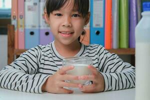 schattig Aziatisch meisje drinken een glas van melk Bij huis in leven kamer. weinig meisje drinken melk in de ochtend- voordat gaan naar school. gezond voedsel in jeugd. foto