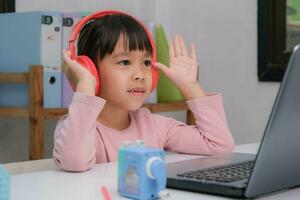 schattig elementair school- meisje vervelend hoofdtelefoons en gebruik makend van een laptop computer. gelukkig Aziatisch kinderen studie online interactief met laptop computer of thuisonderwijs, luisteren naar muziek- of spelen spellen. foto
