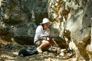 vrouw geoloog gebruik makend van laptop computer onderzoeken natuur, analyseren rotsen of kiezels. onderzoekers verzamelen monsters van biologisch materialen. milieu en ecologie Onderzoek. foto