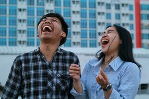 gelukkig Aziatisch paar tiener lachend luid met Open mond en Gesloten oog slijtage gewoontjes overhemd over- stad gebouw achtergrond, op zoek omhoog foto