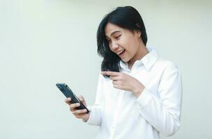 geslaagd speels Aziatisch jong verkoopster verrast op zoek naar smartphone houden Aan handen vervelend wit formeel pak shirt, richten inhoudsopgave vinger naar scherm, staand geïsoleerd foto