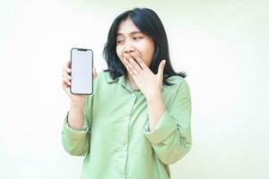 verrast donker haar- Aziatisch vrouw op zoek terzijde tonen smartphone wit scherm en aan het bedekken haar Open mond met handen vervelend groen overmaats overhemd geïsoleerd Aan wit achtergrond foto