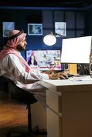 beeld shows een Islamitisch Mens zittend Bij zijn huis kantoor bureau met de bureaublad pc toezicht houden op weergeven een blanco copyspace sjabloon. toegewijd Arabisch vent gebruik makend van de computer hebben een geïsoleerd wit scherm. foto