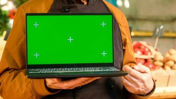 senior verkoper tonen groene scherm Scherm Aan laptop, vervelend schort Bij boeren markt winkel. mannetje verkoper hoding pc met isoated chroma sleutel en blanco copyspace mockup sjabloon. dichtbij omhoog. foto