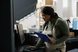 Magazijn leidinggevende op zoek Bij voorraad verslag doen van Aan computer, werken Bij handelswaar kwaliteit controle in magazijn. Afrikaanse Amerikaans werknemer voorbereidingen treffen klanten bestellingen voor Verzending in pakhuis foto