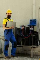 portret van glimlachen goed geïnformeerd ingenieur werken Aan extern hvac systeem, Holding laptop. professioneel draadman optimaliseren condensor prestatie, zorgen voor het opereert Bij maximaal capaciteit foto