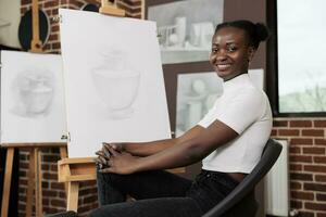 creatief hobby en mentaal Gezondheid. gelukkig vrolijk Afrikaanse Amerikaans millennial meisje hebben pret gedurende kunst klas, zittend Bij ezel en glimlachen Bij camera, uitdrukken haarzelf door tekening foto