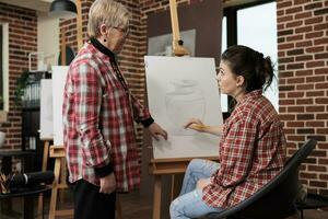 vriendelijk glimlachen senior vrouw leraar staand in de buurt ezel werken met leerling gedurende tekening les. volwassen dochter Bijwonen groep kunst klasse met moeder, besteden tijd samen terwijl aan het leren naar trek foto