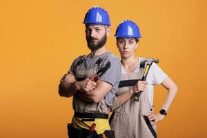 echt bouw arbeiders poseren met paar- van tang en hamer, Holding voorhamer en renoveren gereedschap in studio. Mens en vrouw werken net zo constructeurs met overall en helm. foto