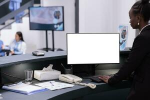 Afrikaanse Amerikaans receptioniste gebruik makend van wit geïsoleerd scherm Aan computer terwijl werken Bij ziekenhuis registratie balie. ontvangst arbeider op zoek Bij mockup sjabloon gedurende controle bezoek afspraak foto