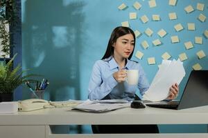 professioneel Aziatisch vrouw drinken een kop van heet koffie in kantoor terwijl herzien maandelijks statistieken rapporten. vrouw bedrijf persoon behandeling papierwerk voordat beheer ontmoeting. foto