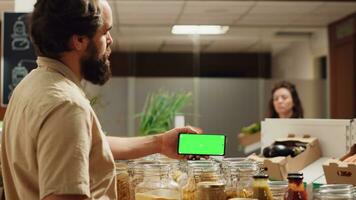 Mens gebruik makend van groen scherm landschap mode smartphone in eco vriendelijk supermarkt. cliënt in lokaal kruidenier winkel toepassingen chroma sleutel telefoon terwijl boodschappen doen voor biologisch groenten, pannen schot foto