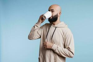 jong Arabisch Mens drinken koffie van wit kop en op zoek Bij camera. Arabisch kaal gebaard persoon vervelend gewoontjes kleren nippen thee, genieten van heet drank van mok studio portret foto