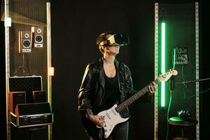 vrouw performer spelen zwaar metaal lied gebruik makend van elektrisch gitaar terwijl vervelend virtueel realiteit stofbril gedurende muziek- sessie in geluid studio. performer met vr koptelefoon hebben grunge concert simulatie foto