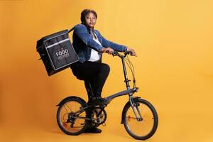 voedsel levering werknemer rijden fiets terwijl draag- meenemen thermisch rugzak klaar naar leveren bestellen naar cliënt gedurende lunch tijd, staand over- geel achtergrond. voedsel onderhoud en vervoer foto