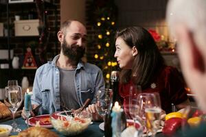 partners vieren Kerstmis vakantie met vrienden, acteren zoet Bij avondeten tafel met familie. verschillend mensen genieten van bijeenkomst met eigengemaakt maaltijd en wijn naar vieren Kerstmis vooravond, winter seizoen. foto