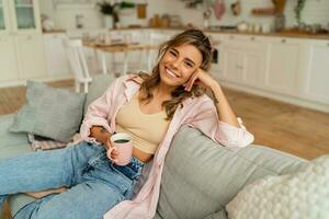 lief vrouw in gewoontjes kleren resting un sofa in knus leven kamer. Holding kop van cappuccino. foto