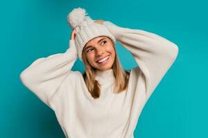 studio winter portret van glimlachen blond vrouw in wit hoed en whoo trui poseren in studio over- turkoois achtergrond. verkoudheid seizoen accessoires en mode. foto